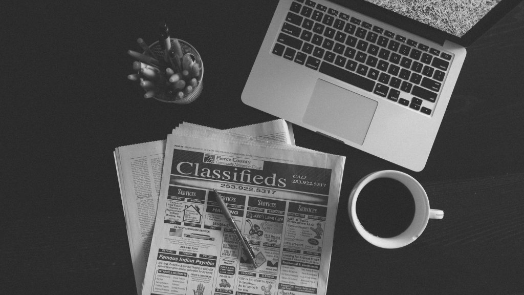A black and white photo of an open laptop and the classifieds section of a newspaper